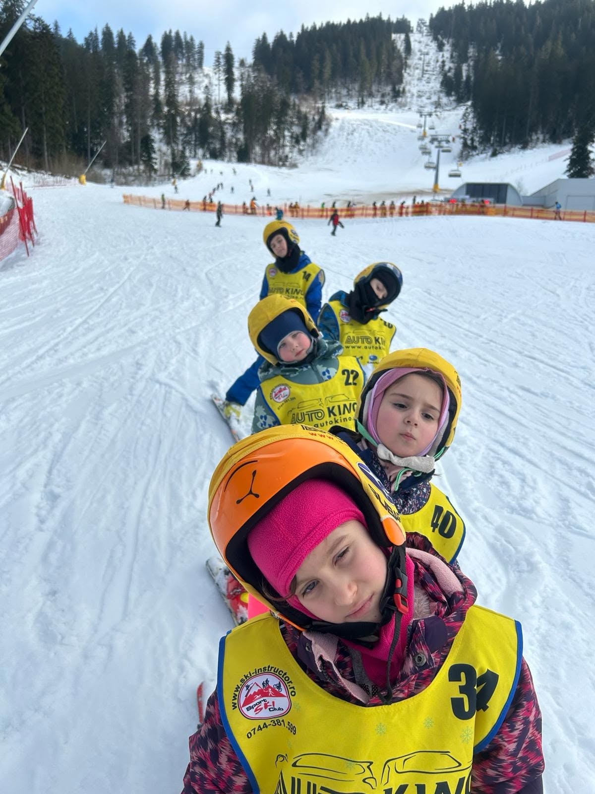 Copii cu cască și veste numerotate pe pârtie
